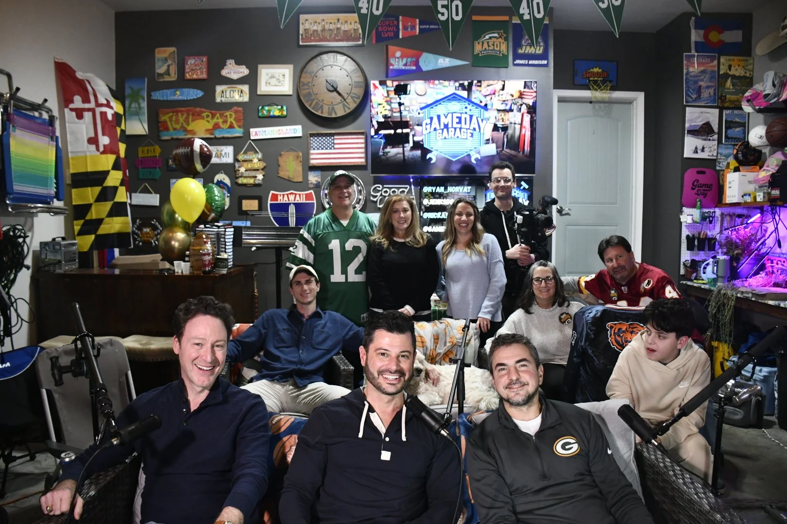 A group of people gathered in a decorated room with sports memorabilia and banners, some holding microphones and filming equipment, indicating a podcast or recording session.