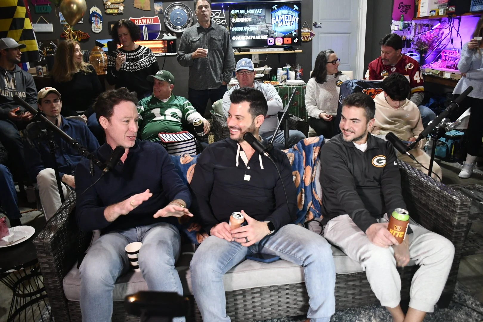 Group of people gathered in a room, socializing and enjoying drinks, with microphones set up, indicating a podcast or casual discussion setting.