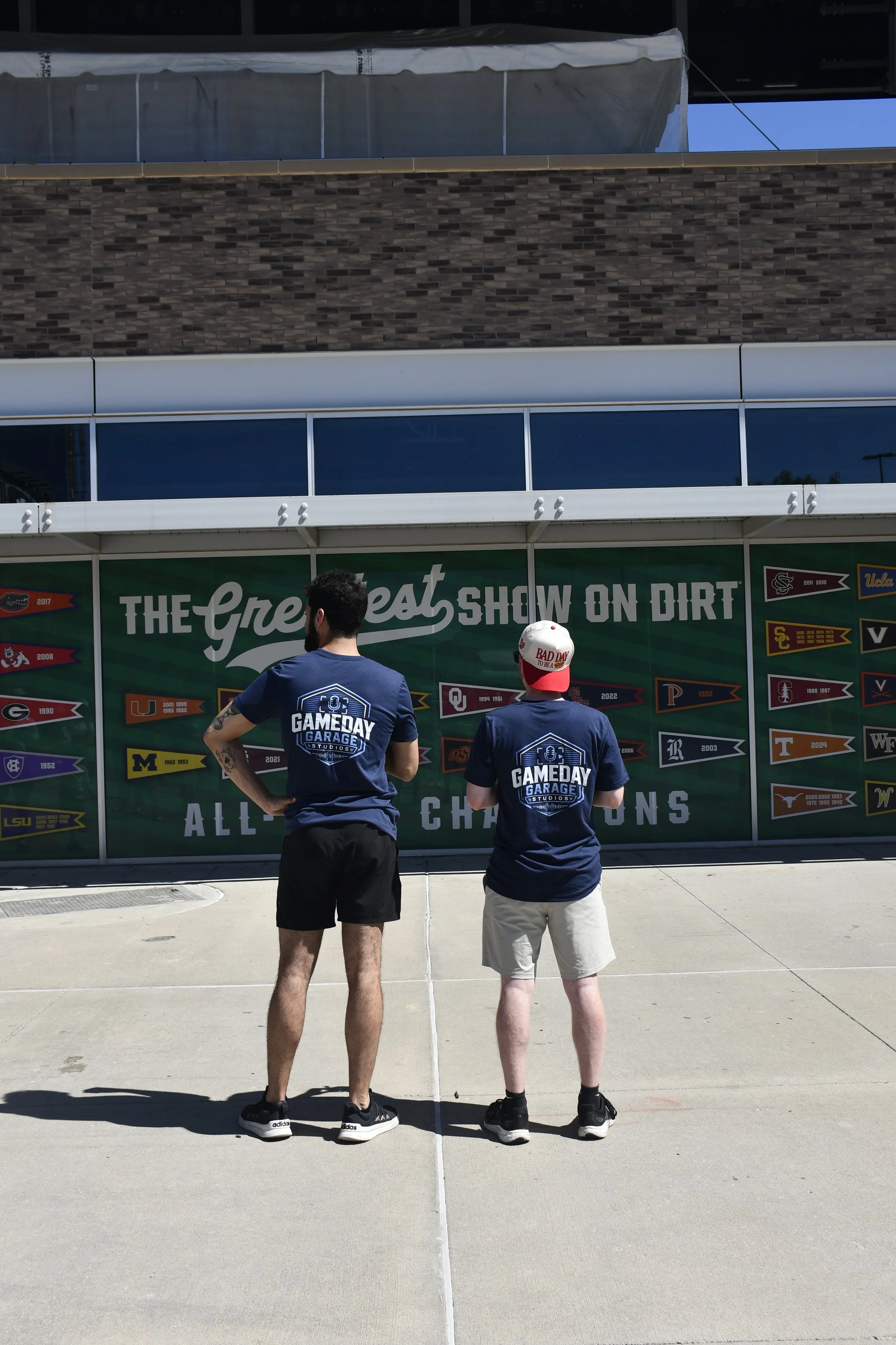 Two men standing on sidewalk with their backs to the camera, watching an event banner for college football teams and championships, with a building behind them.