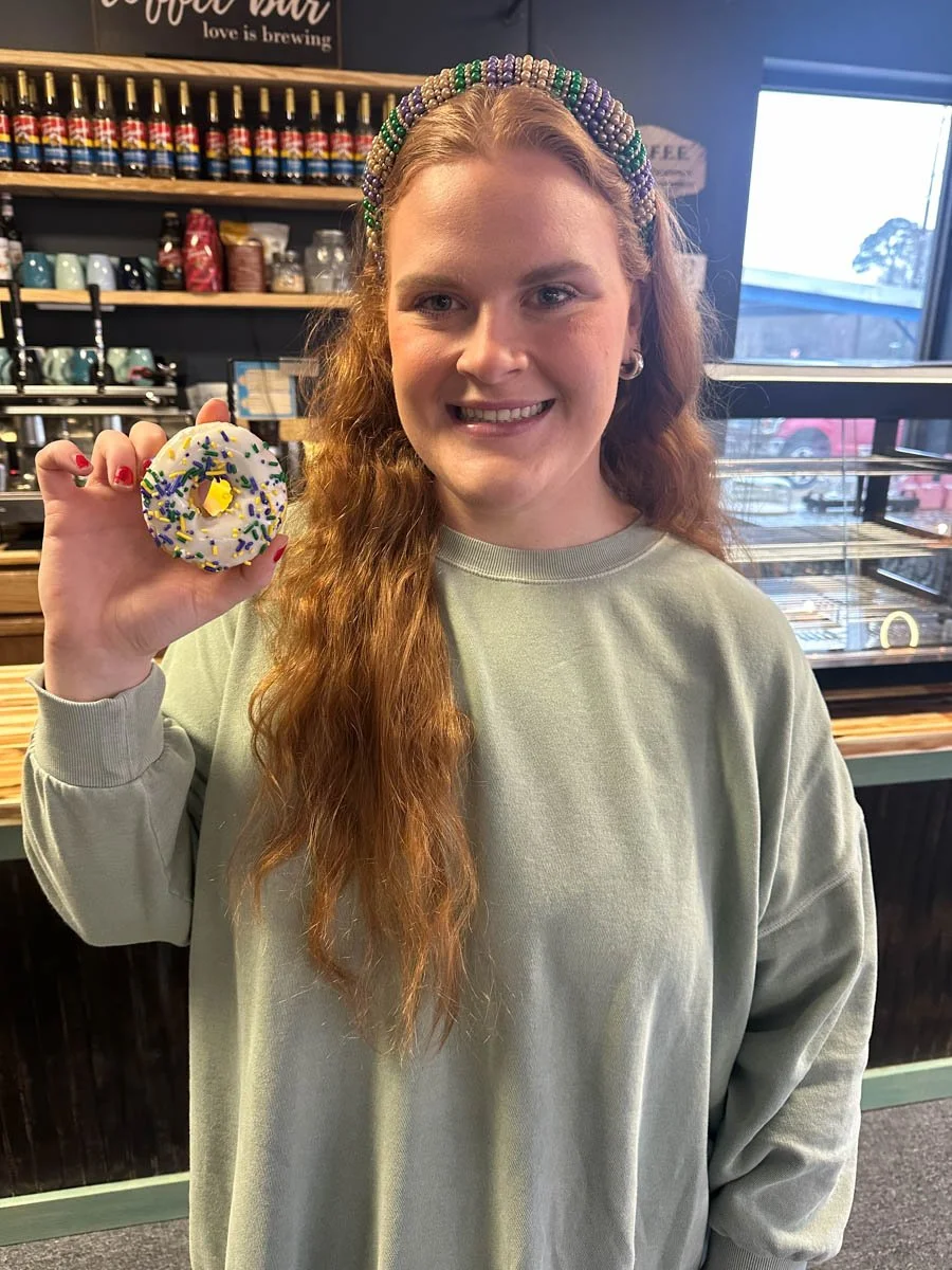 A young woman with long, curly red hair holding a decorated donuts in a cafe. She is smiling and wearing a dotted headband and a light green sweatshirt. Behind her, there are shelves with jars and bottles, and a window showing parked cars outside.