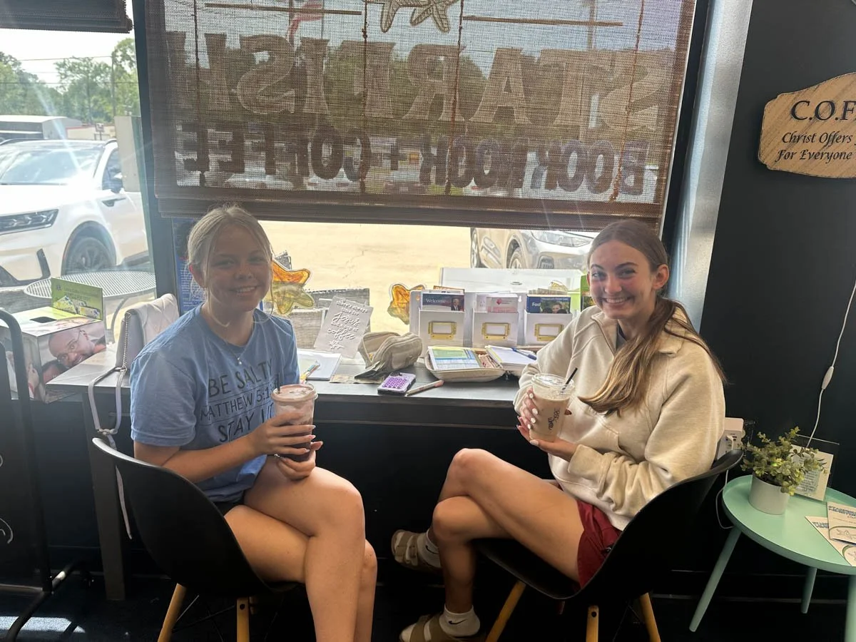 Two young women sitting by a window in a coffee shop, smiling and holding drinks, with a parking lot outside.