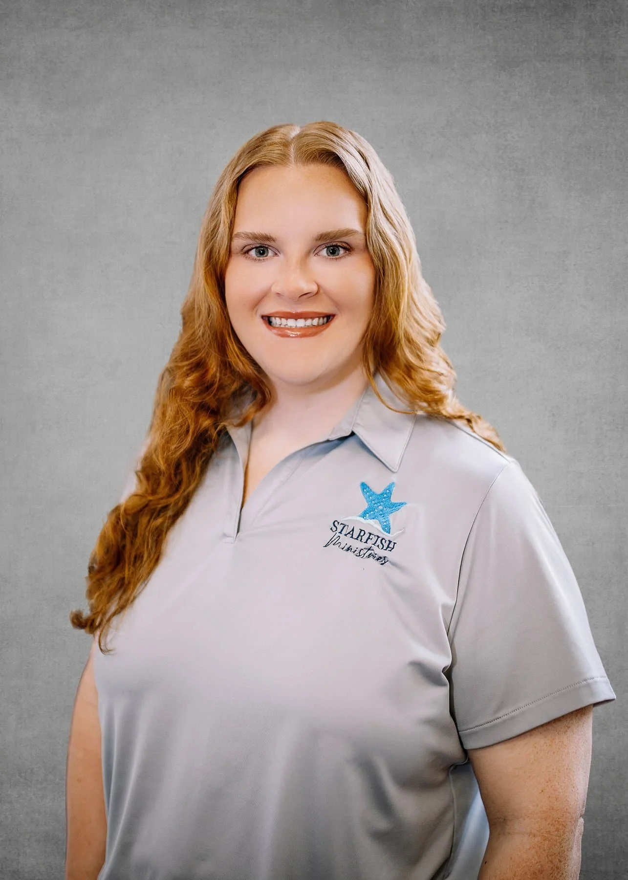 A woman with long, curly red hair wearing a light gray polo shirt with a starfish logo and the text 'Starfish' and 'Damanhy' embroidered on it, standing against a plain gray background.