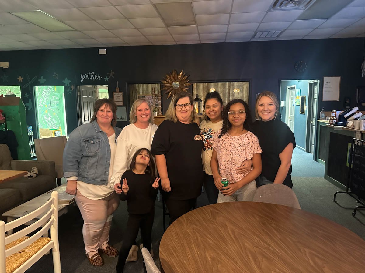 Group of six women and one young girl smiling and posing together inside a restaurant or cafe. The background includes dark walls, a clock, and decor.