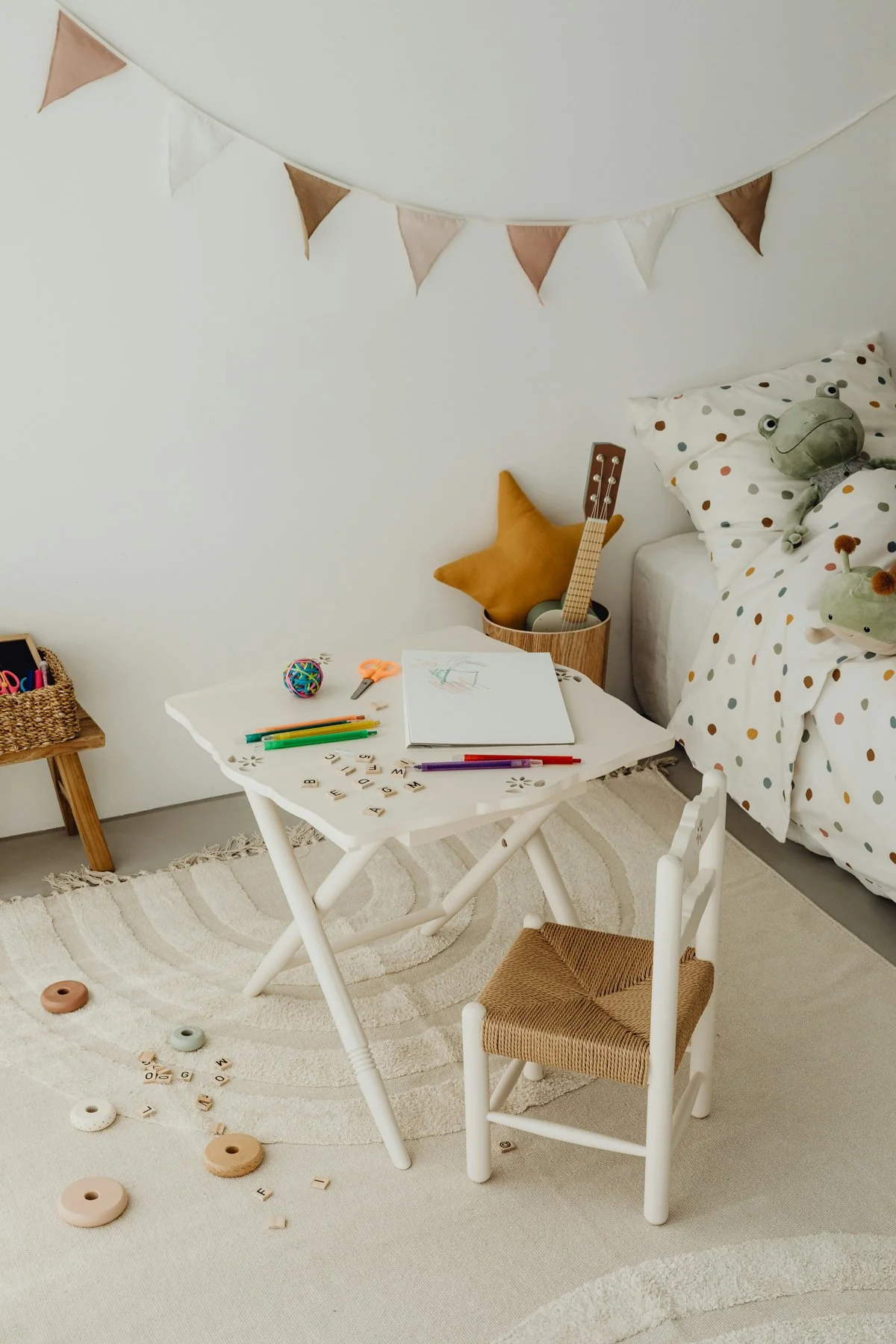 A cozy children's bedroom with a white table, scattered alphabet tiles, drawing supplies, and toys including plush frogs and a star-shaped cushion. There is a bed with polka dot bedding and stuffed frogs, a small woven basket with art supplies, a star-shaped cushion, and a guitar with a string holder in a wooden container. A string of triangular fabric bunting decorates the wall.
