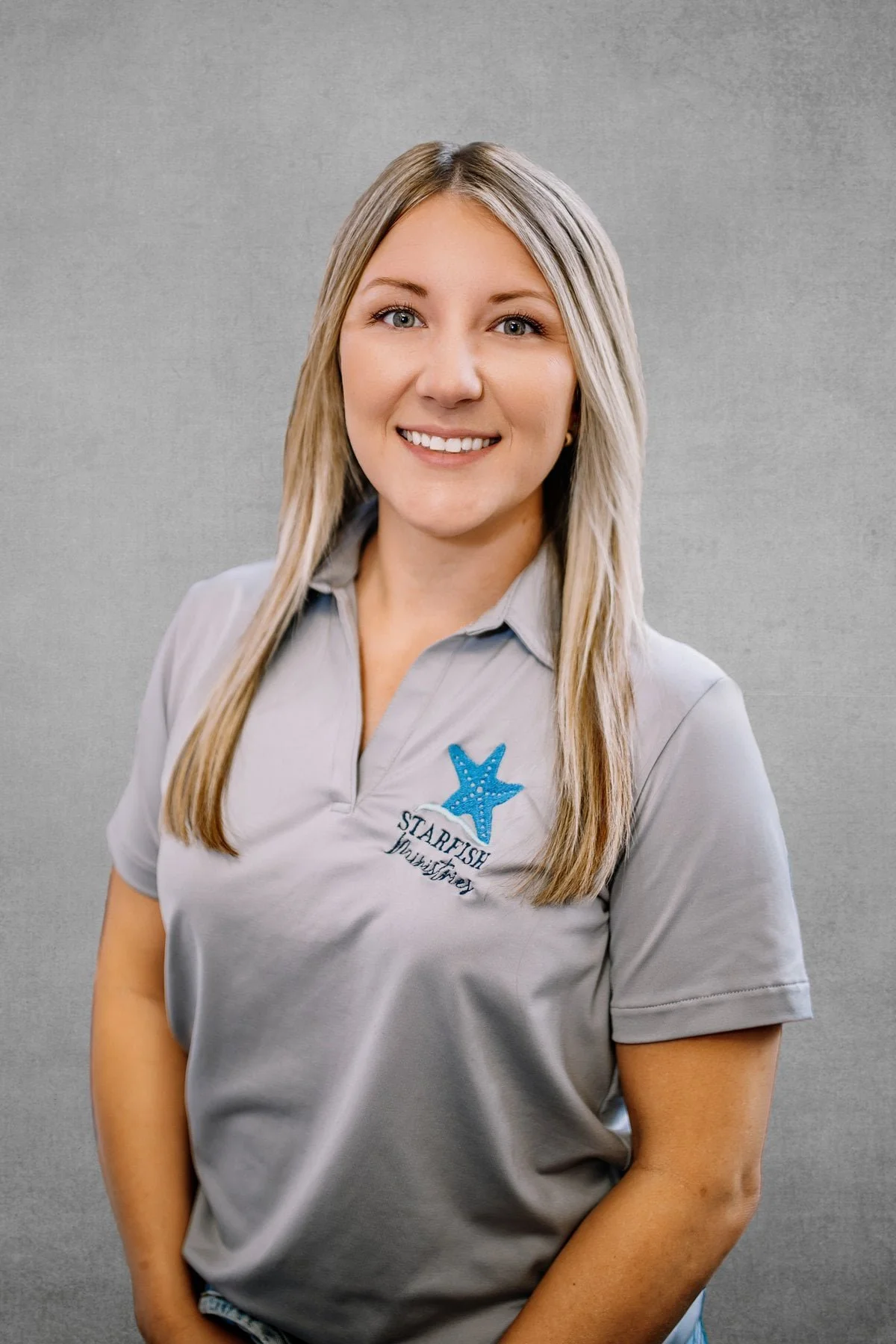 A woman with long blonde hair smiling, wearing a light gray polo shirt with a 'Starfish Destin' logo featuring a blue starfish embroidered on the left side, posed against a plain gray background.