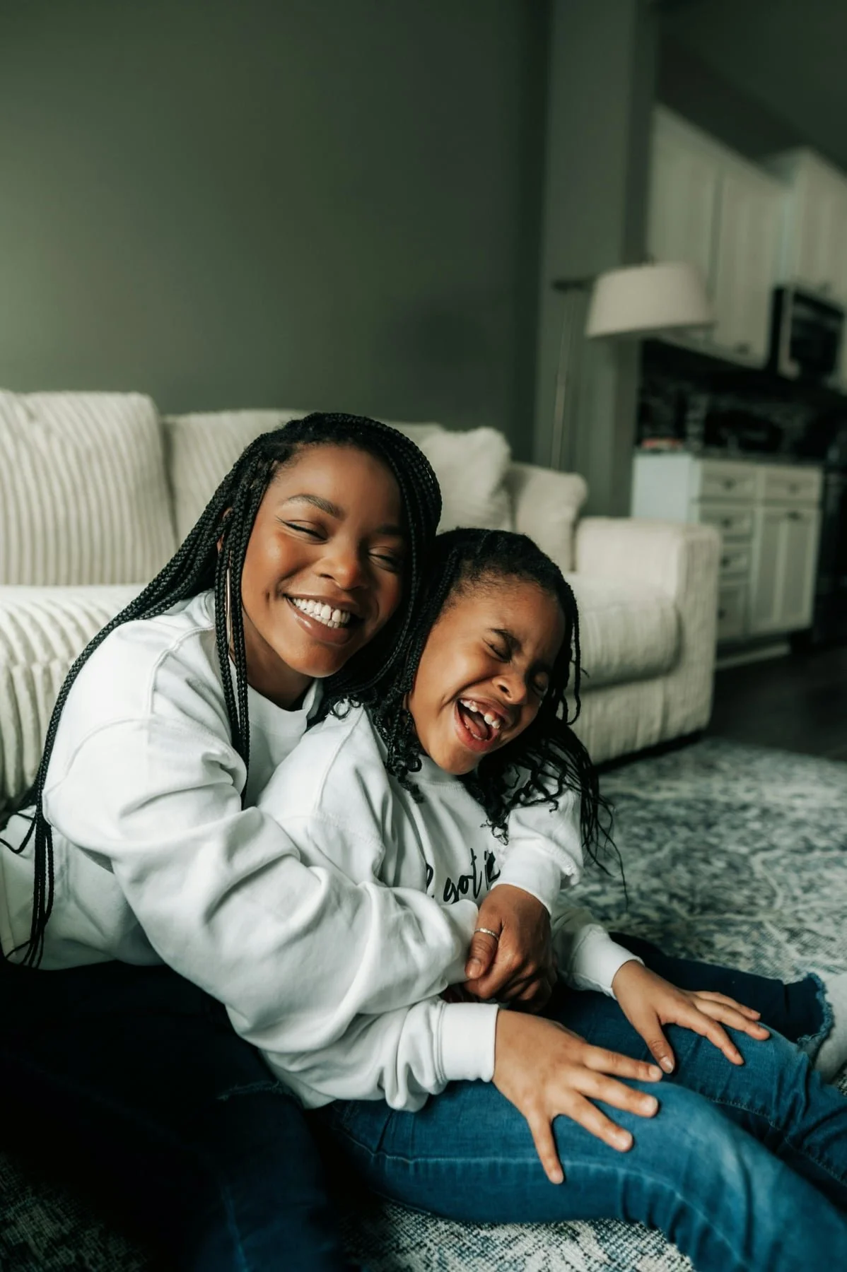 A woman and a young girl sitting on the floor together, both smiling and laughing joyfully, in a cozy living room with a striped sofa and a rug in the background.