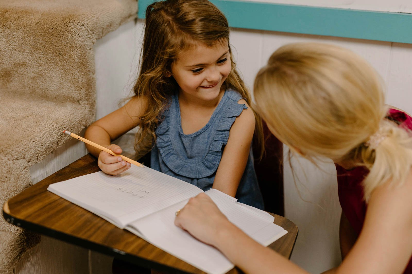A young girl sitting at a small table, smiling and holding a pencil, as another person leans in and engages with her, with an open notebook in front of them.