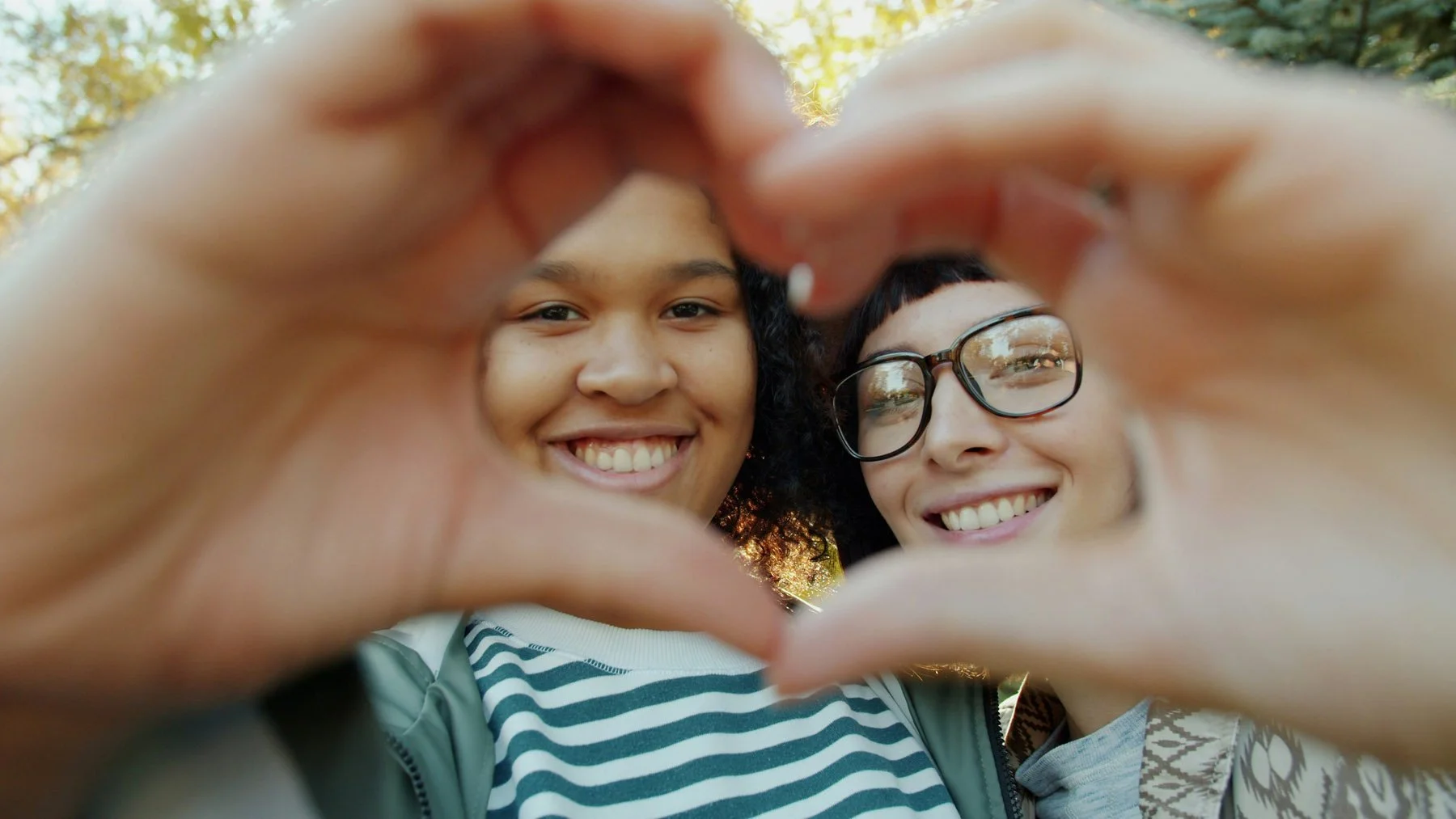 Two women smiling and looking at the camera while forming a heart shape with their hands. They are outdoors with trees and sunlight in the background.