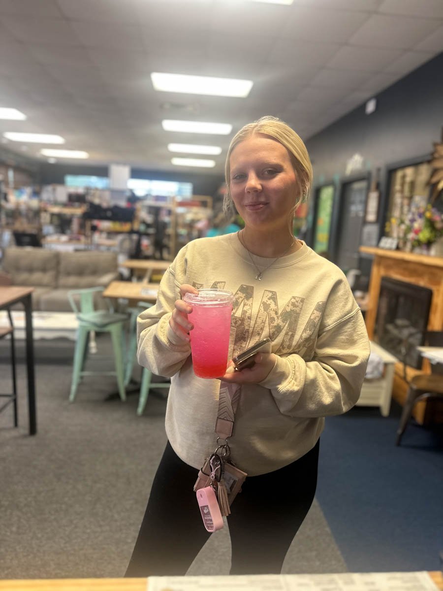 A woman holding a pink beverage in a glass jar, standing inside a store or cafe with various furniture and decor in the background.