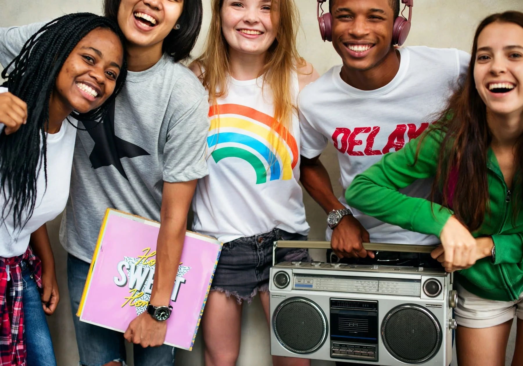 Group of five young people smiling and having fun together, with one holding a boombox and a woman holding a pink sign that says 'The Sweet T.'