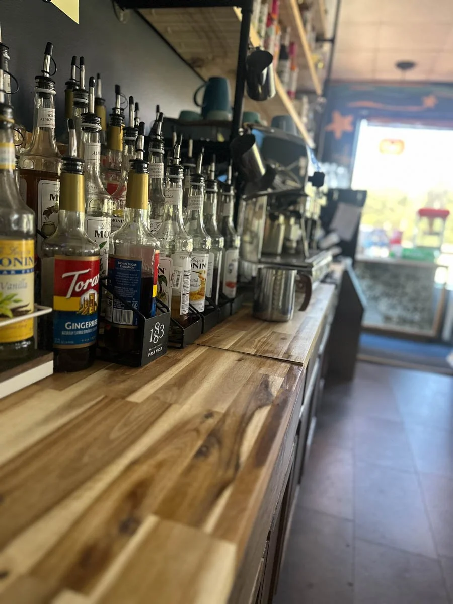 A wooden countertop in a café or restaurant with spice and syrup bottles lined up, coffee cups, and brewing equipment in the background, with a door and window showing outdoor sunlight.