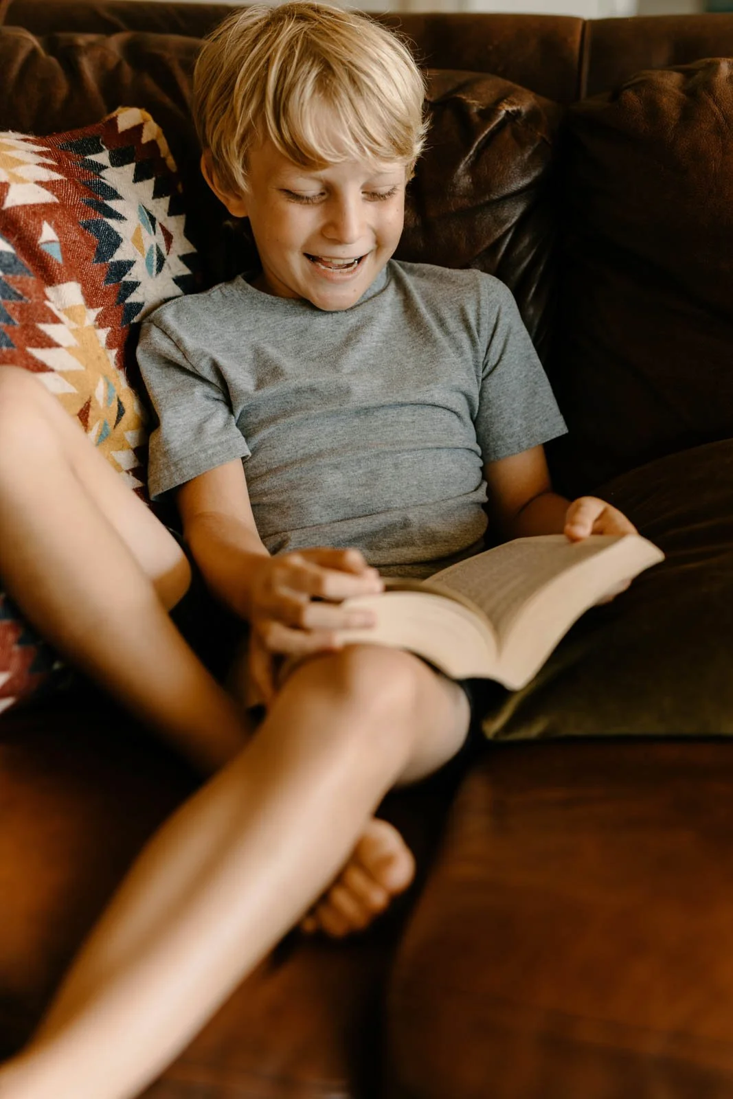 A young boy with blonde hair smiling and looking at a book while sitting on a dark brown leather couch, with a patterned pillow behind him.