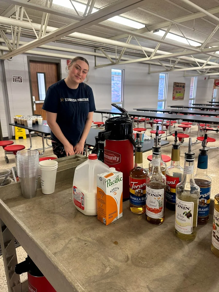 A young woman with a ponytail wearing a navy t-shirt standing behind a counter with various syrups and drinks, in a cafeteria or dining hall with red stools and long tables in the background.