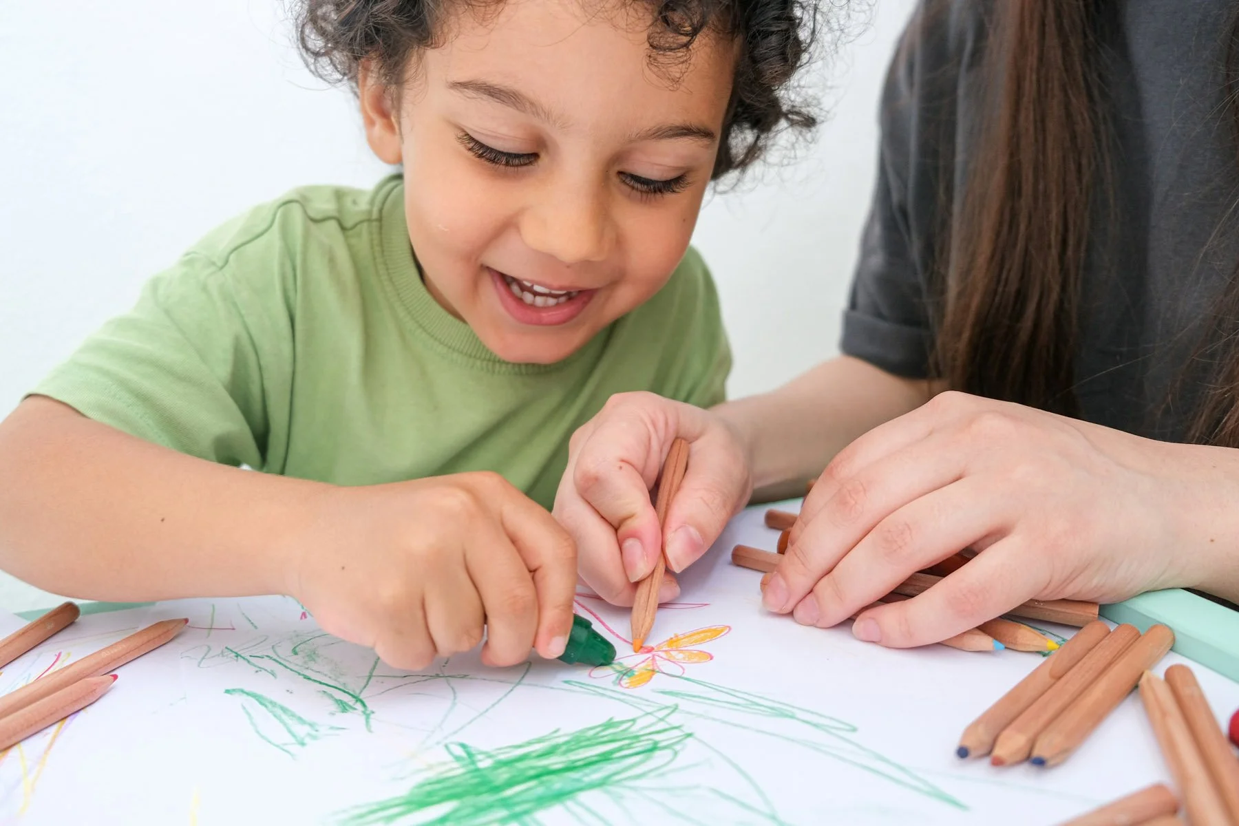 A young boy and an adult drawing together on a large sheet of paper using colored pencils, with scattered pencils around them.