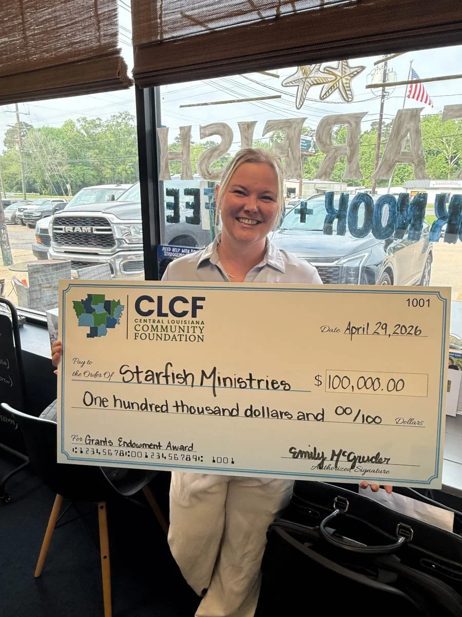 A woman standing inside a building, holding a large check made out to Starfish Ministries for $100,000. The check is from the Central Louisiana Community Foundation, dated April 29, 2026, and signed by Emily McGuder. The background shows a window wit