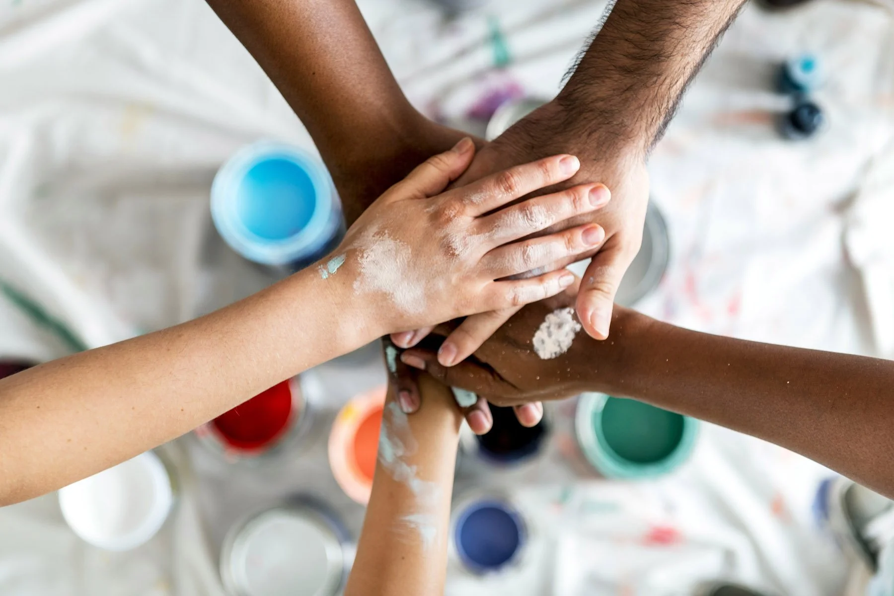 Multiple hands stacking together, with the background showing open paint cans in various colors.