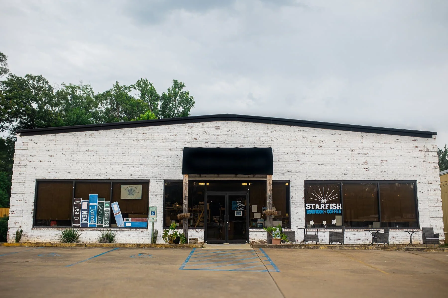 Front view of a white brick building with large glass windows, a black awning over the entrance, and a sign that reads 'Starfish Bookstore + Coffee.' There are parking spaces in front, some with blue markings, and a few potted plants near the entrance. Tall trees are visible behind the building under a cloudy sky.