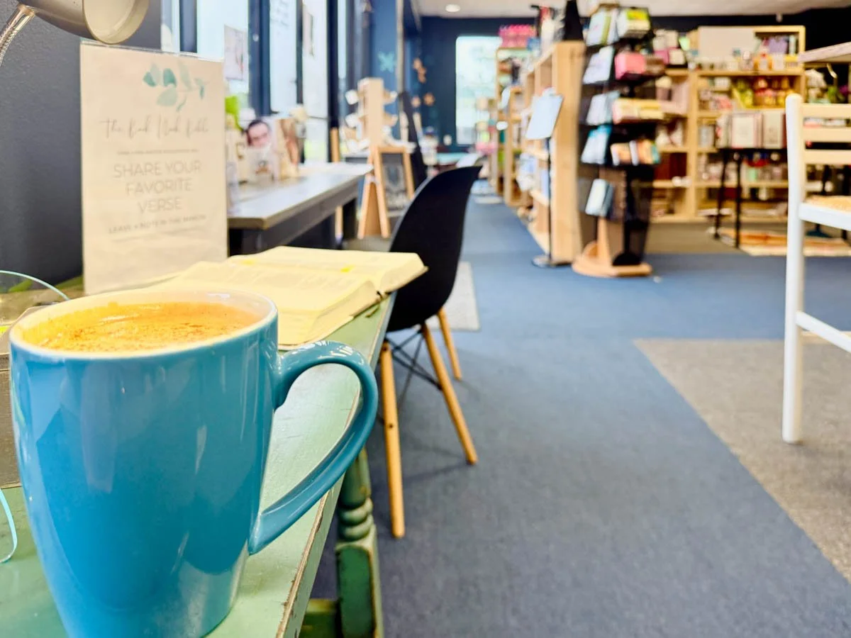A blue mug filled with a hot beverage on a table inside a bookstore or cafe, with bookshelves and chairs in the background.