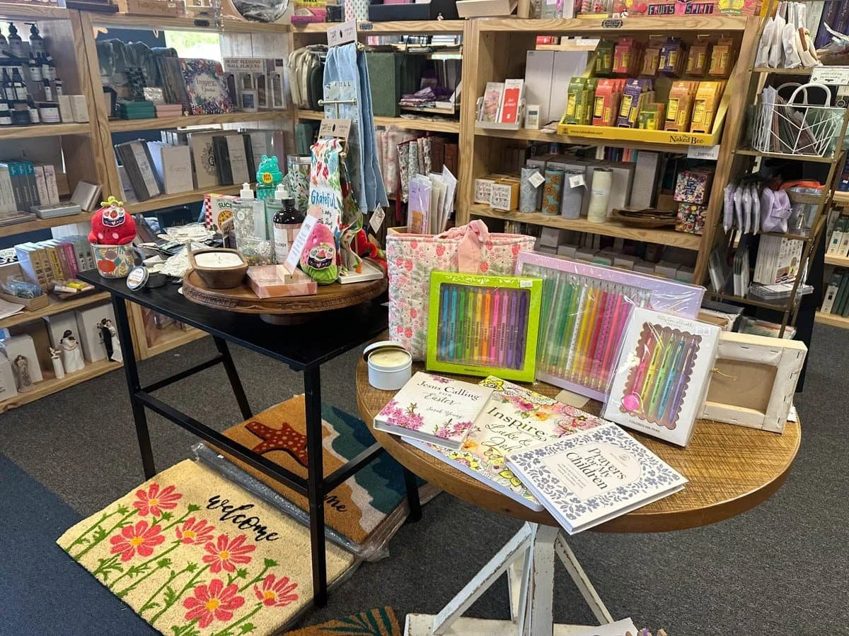 Display of greeting cards, colorful pens, and notebooks on round tables inside a bookstore or gift shop with shelves of books and home décor items in the background.