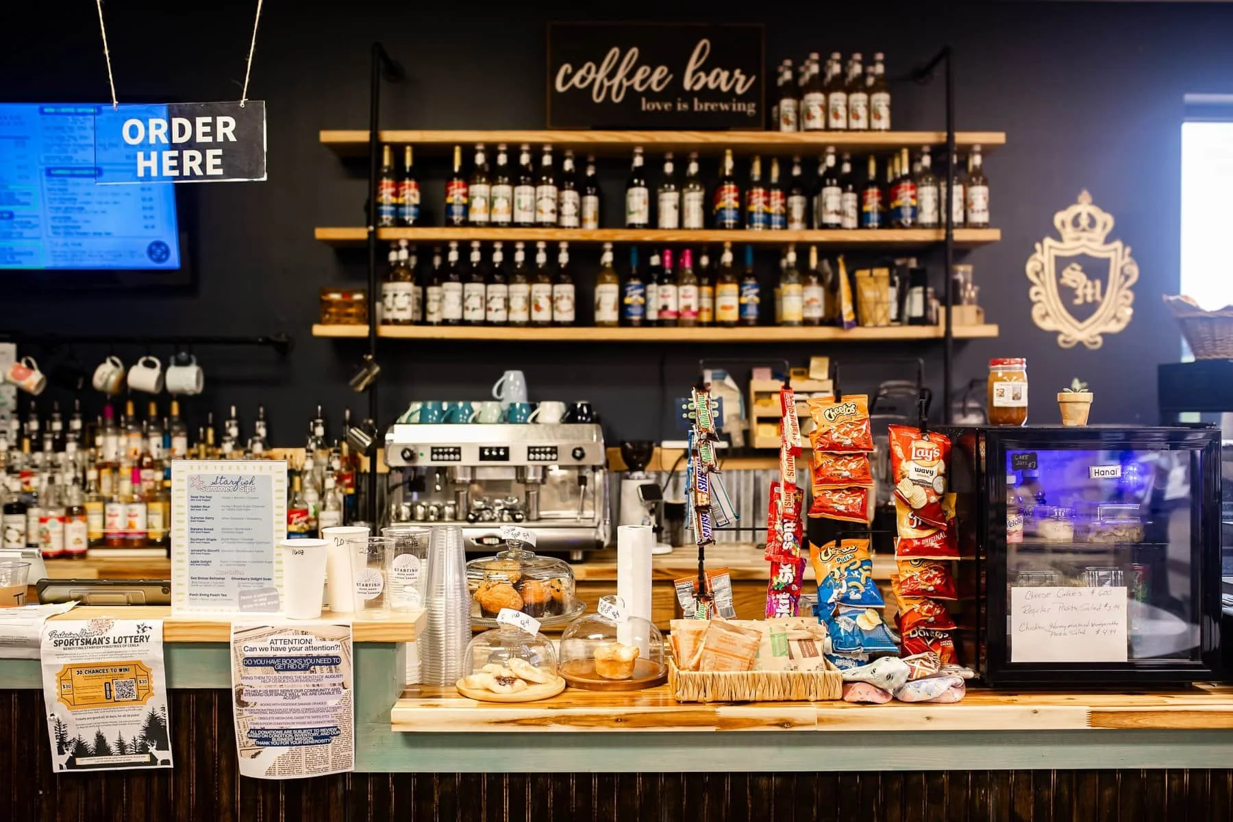A coffee shop counter with snacks, coffee cups, a coffee machine, and shelves with bottles of syrup or flavoring. There is a sign that says 'ORDER HERE' and a menu board. Snacks like chips and baked goods are displayed on the counter. A refrigerator contains cheesecake slices labeled 'Hana' and other items.