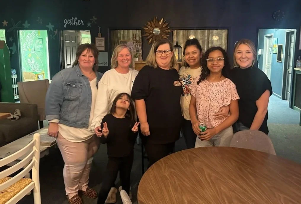 Group of seven women and a young girl posing in a cozy indoor space with dark walls, decorative mirror, and festive wall art.