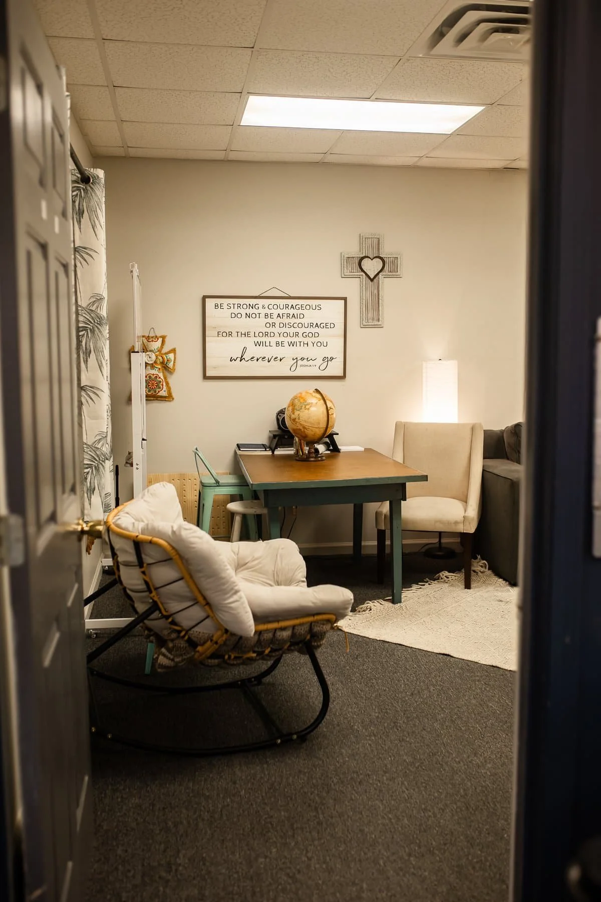 A cozy office space viewed through a doorway. It features a wooden desk with a globe on top, surrounded by various chairs including a light-colored armchair, a green chair, and a cream-colored high-back chair. The wall has a cross with a heart and a framed inspirational quote that reads, "Be strong & courageous do not be afraid or discouraged for the Lord your god will be with you wherever you go." There is a small lamp emitting warm light, a rug on the dark carpeted floor, and decor including a curtain with a nature motif and wall hanging.
