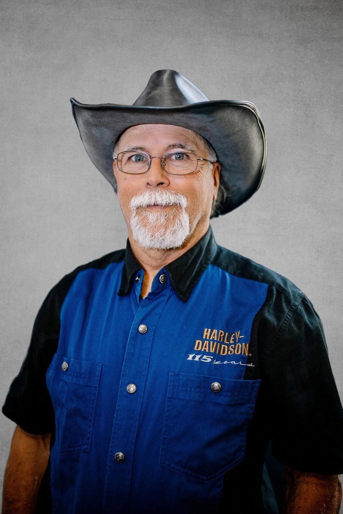 A man with glasses and a white beard wearing a black cowboy hat and a black and blue Harley-Davidson shirt.