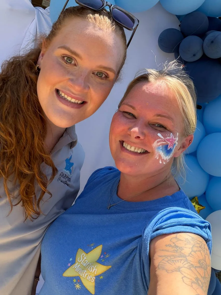 Two smiling women taking a selfie against a backdrop of blue balloons. One woman has long curly red hair, sunglasses on her head, and is wearing a light gray shirt. The other woman has blonde hair, a face painting of a pink star and blue heart on her cheek, and is wearing a blue T-shirt with a yellow star and the text "Starfish Ministries."