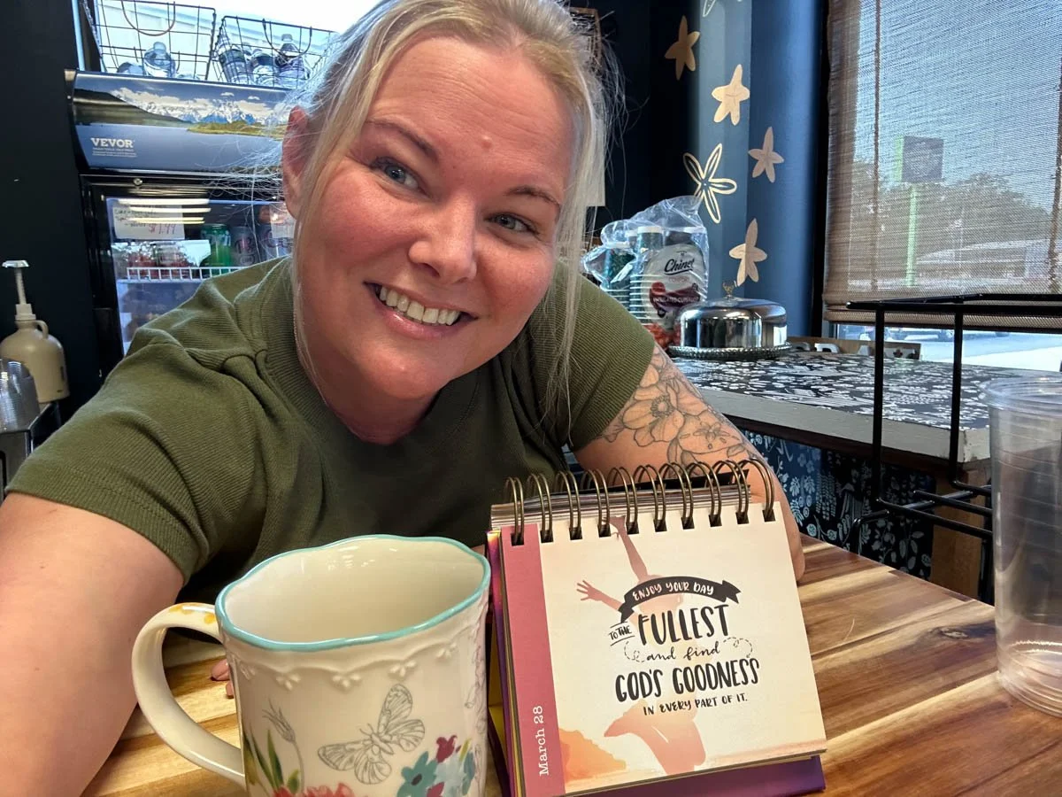 A smiling woman with blonde hair and a tattoo on her arm, sitting at a table in a café. There is a floral mug and a colorful motivational card on the table. Behind her, there is a refrigerator, bottled water, and a window with curtains.