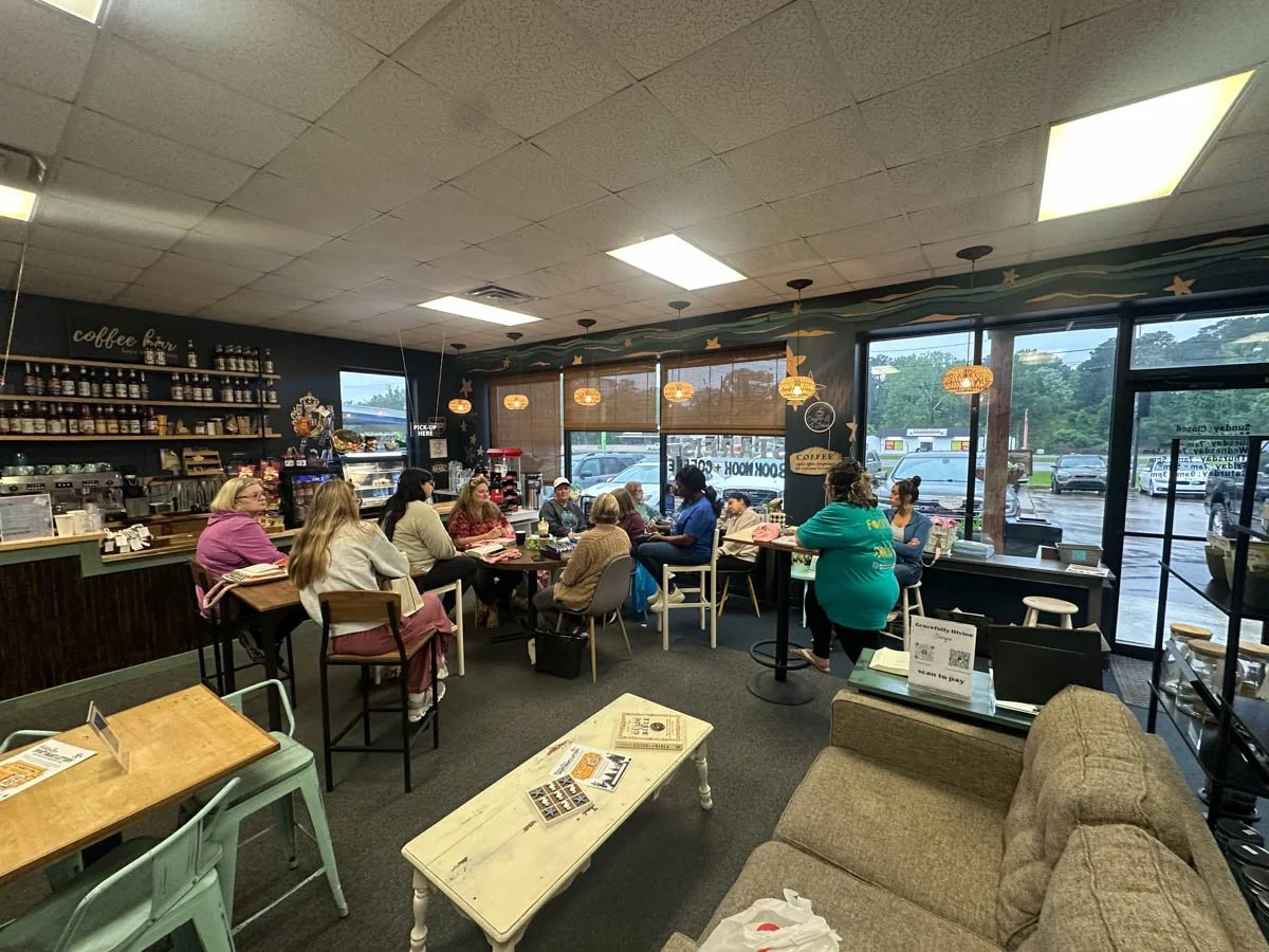 Inside a coffee shop with a group of people sitting and chatting around tables, with a barista counter, coffee jars, and large windows showing a parking lot outside.