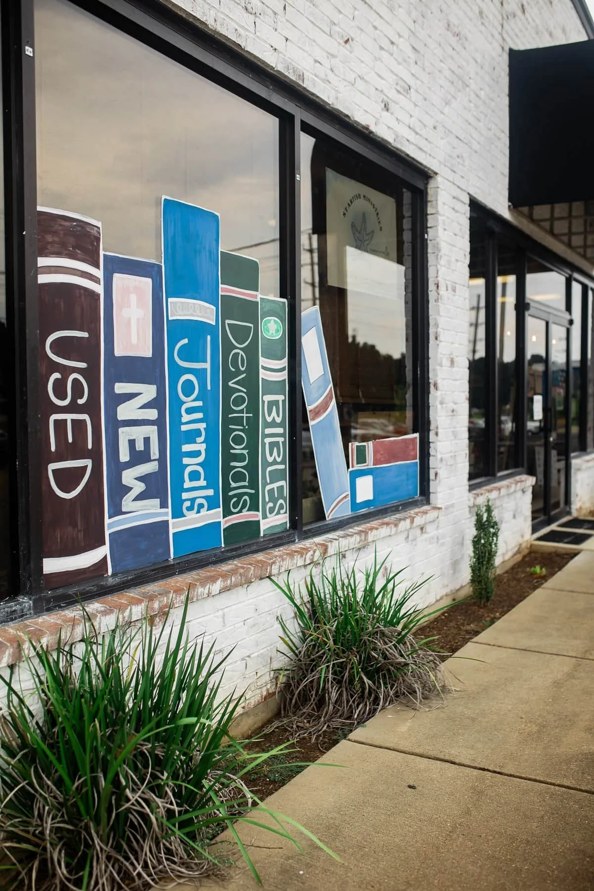 Storefront window with painted book spines labeled Old Testament, New Testament, Devils, and Psalm, with grass plants and a sidewalk in front of the white brick building.
