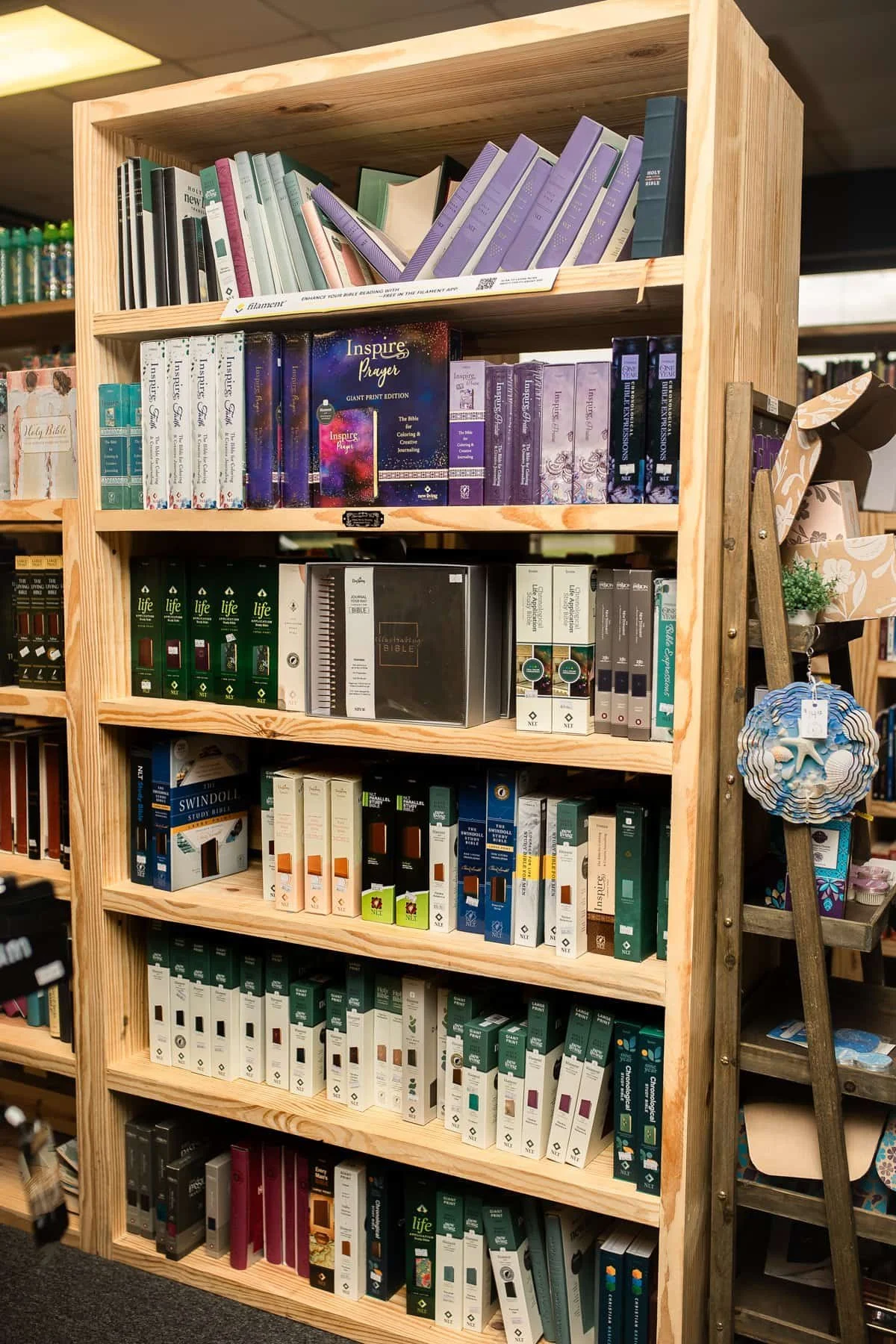 Wooden bookshelf filled with religious and inspirational books, with a ladder leaning against it and a small plant in a decorative pot on the side.