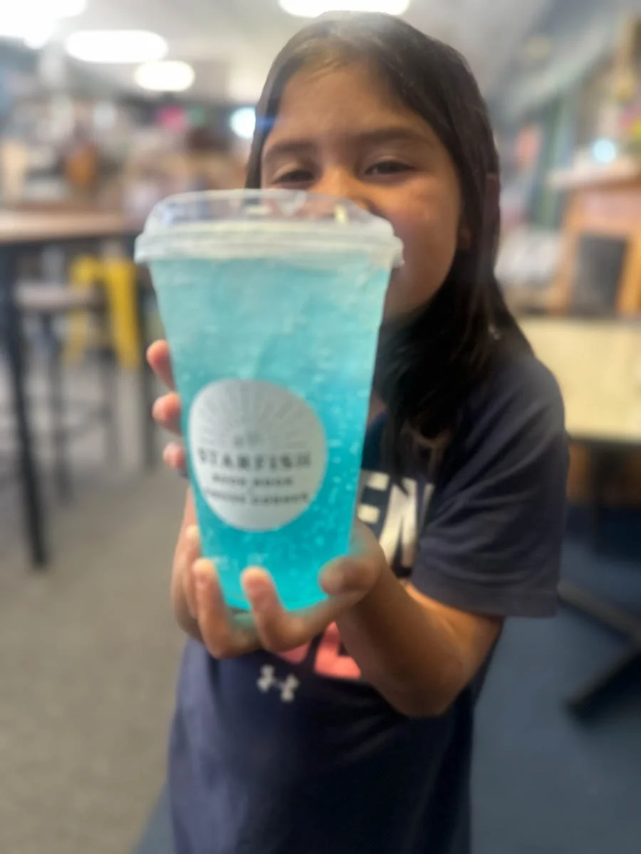 A young girl in a dark blue shirt holding a large cup of blue slushy drink at a cafe or restaurant.