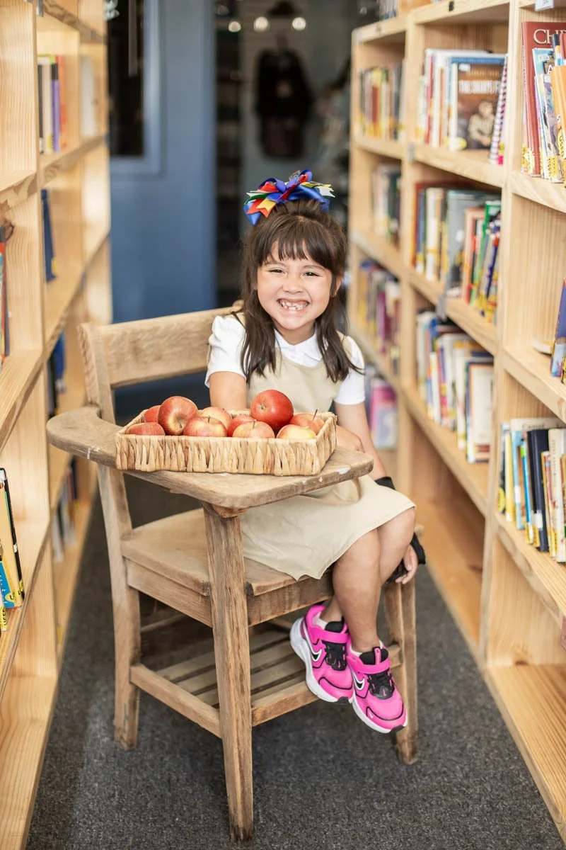 Young girl sitting in a wooden chair in a bookstore, holding a tray of apples, smiling with braces, wearing pink sneakers and a colorful bow in her hair.