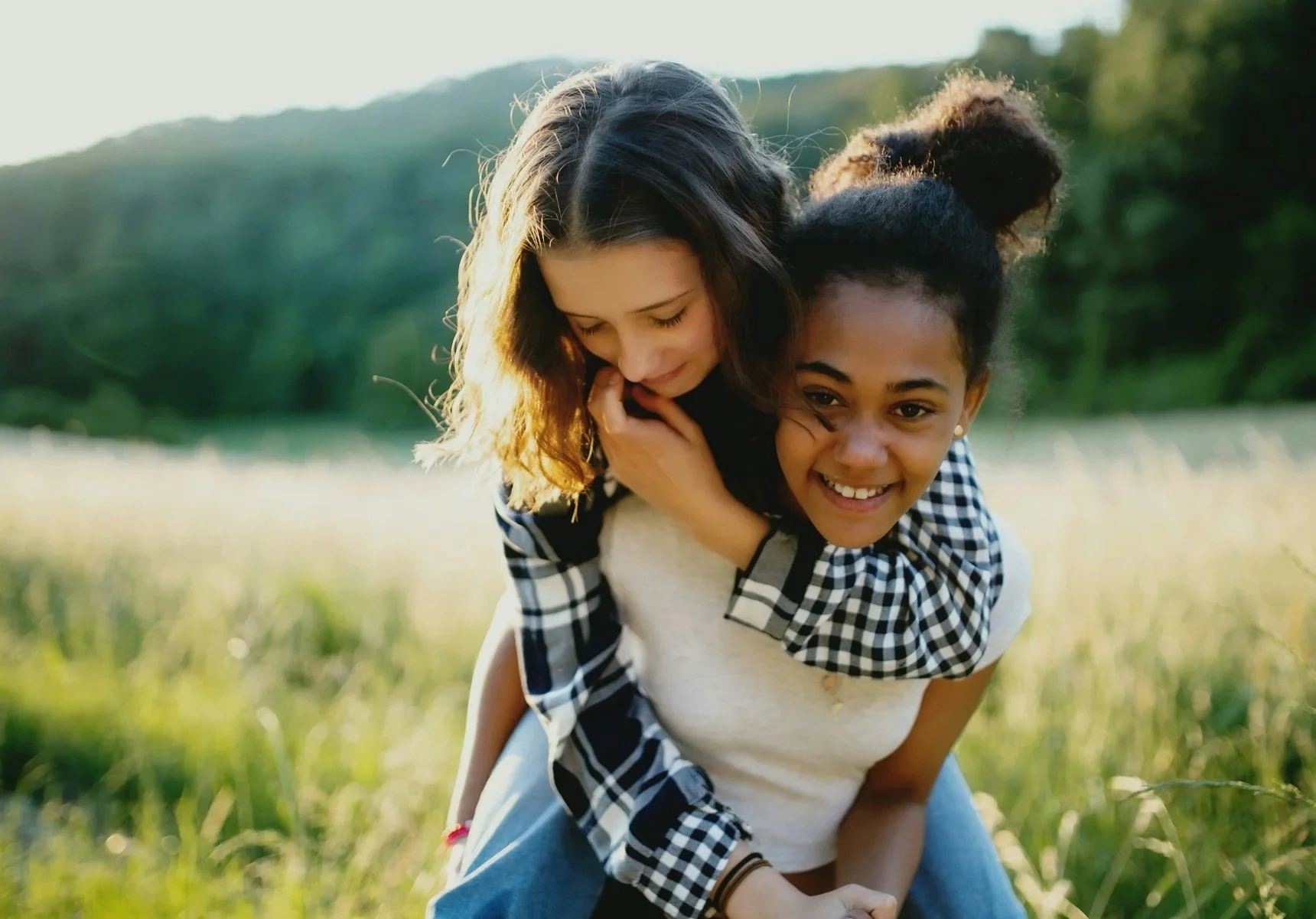Two women playing outside in a grassy field with mountains in the background. One woman is giving the other a piggyback ride. Both are smiling and enjoying the moment.