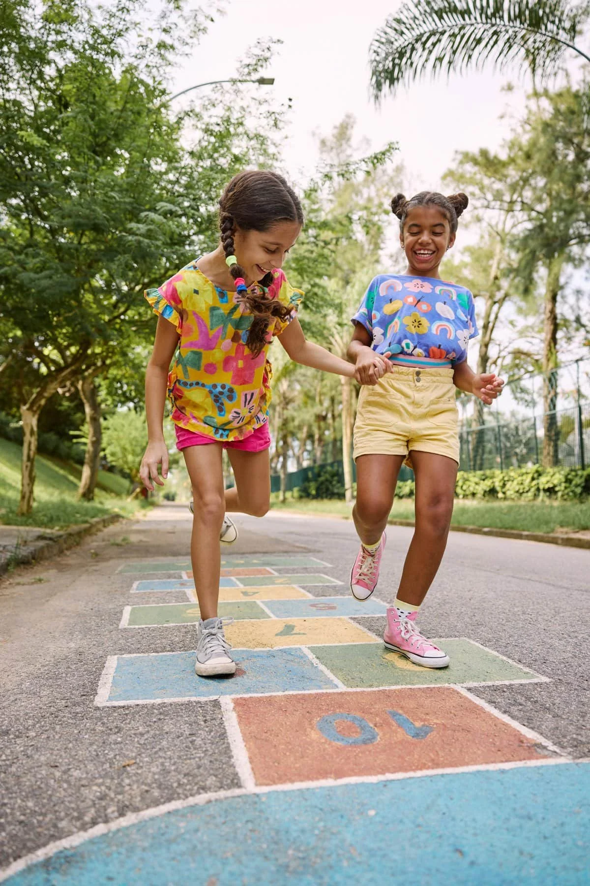 Two young girls with braided hair, wearing colorful t-shirts and shorts, playing hopscotch on a paved path in a park with green trees and a fence in the background on a bright day.