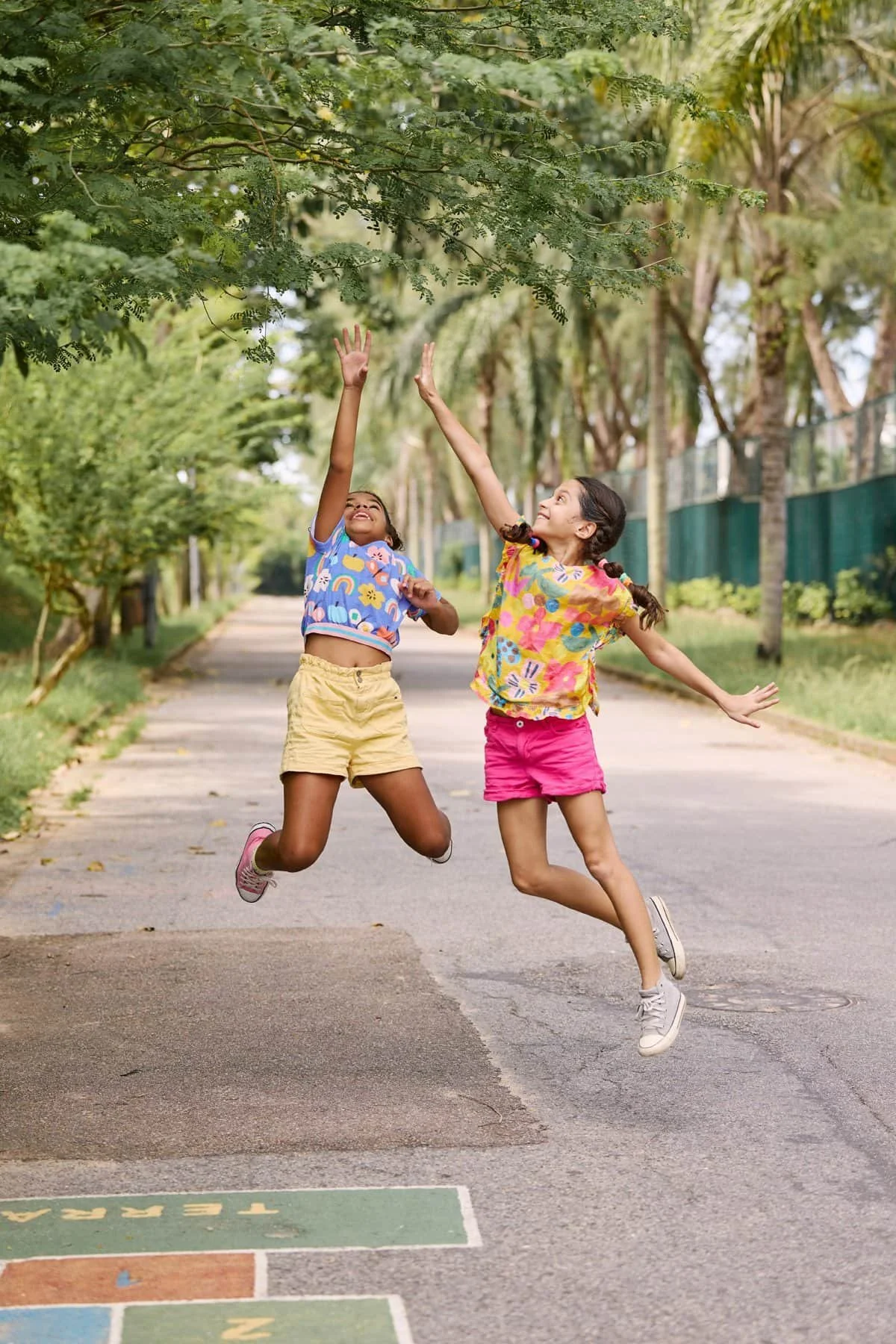 Two young girls jumping and playing on a sidewalk in a park, reaching towards the trees above them.