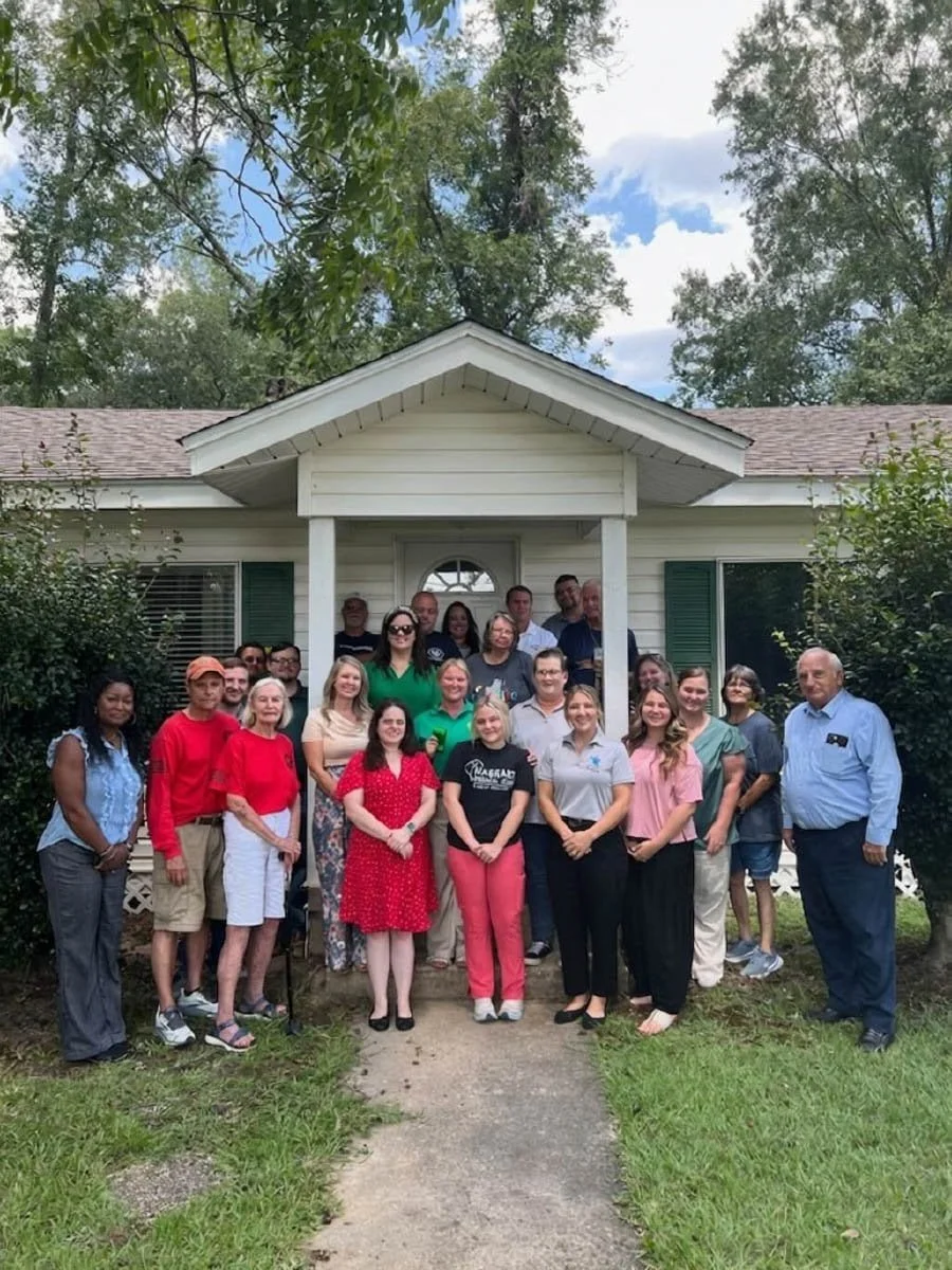 Group of people standing outside a house on a sidewalk, with trees and bushes in the background.