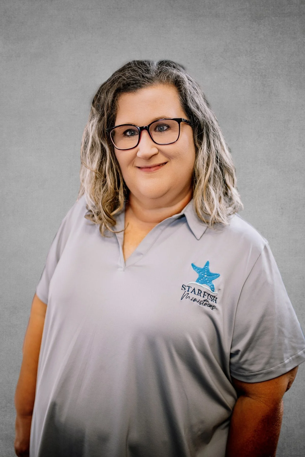 A woman with shoulder-length wavy gray hair and glasses, wearing a gray polo shirt with an embroidered logo of a blue starfish and the text 'Star Fish Vina Del Mar'. She is smiling slightly in front of a neutral gray background.