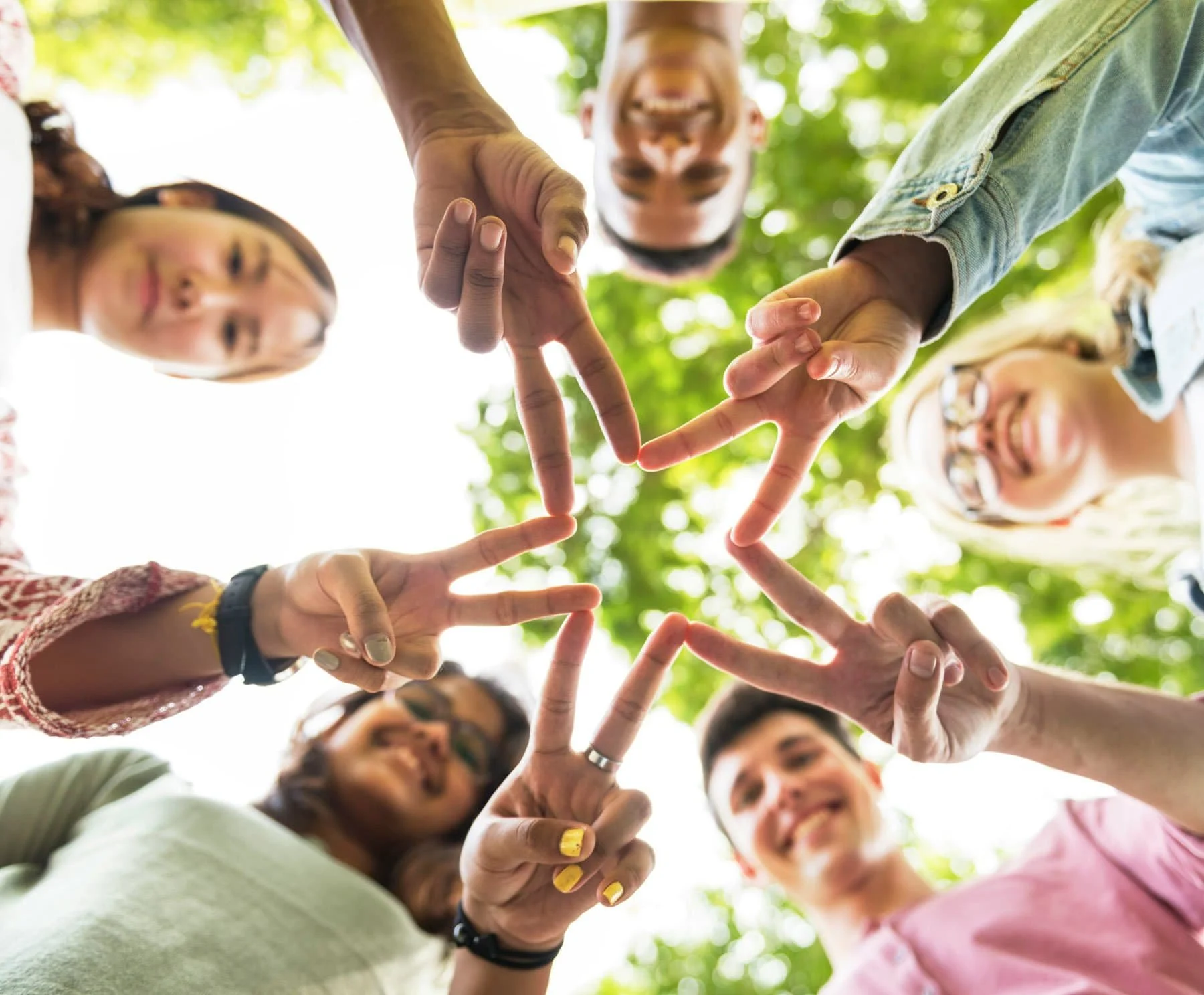 A group of diverse smiling people standing in a circle outdoors with a bright green leafy background, reaching inwards with their hands to form a star shape in the center.