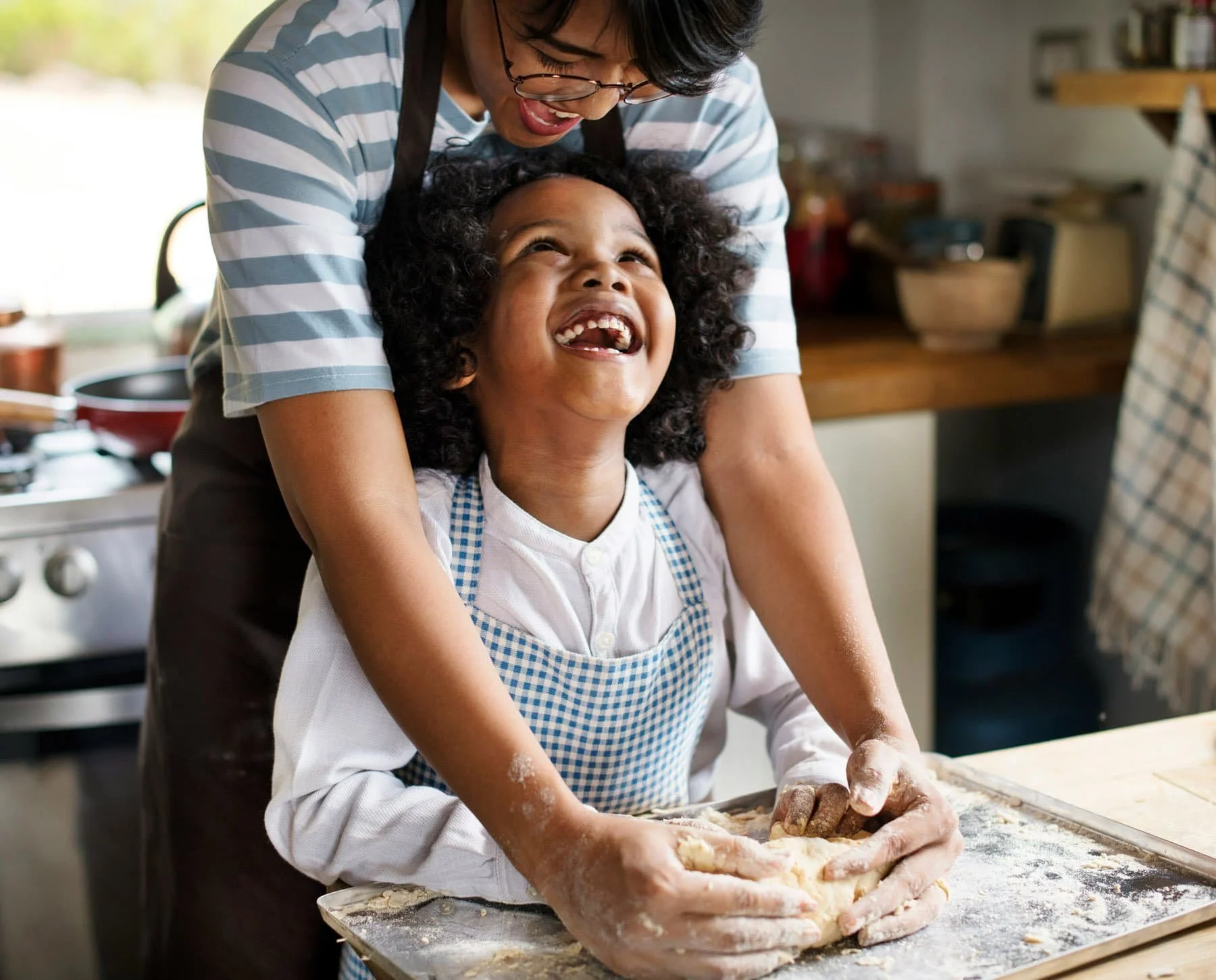 A woman and a girl are baking together in a kitchen, with the girl smiling and looking up at the woman.