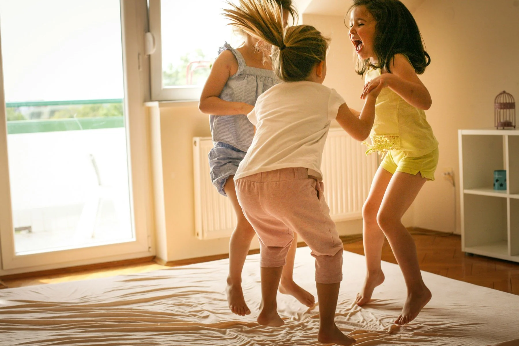 Four children playing and laughing together on a bed in a sunlit room with large windows and a balcony door.