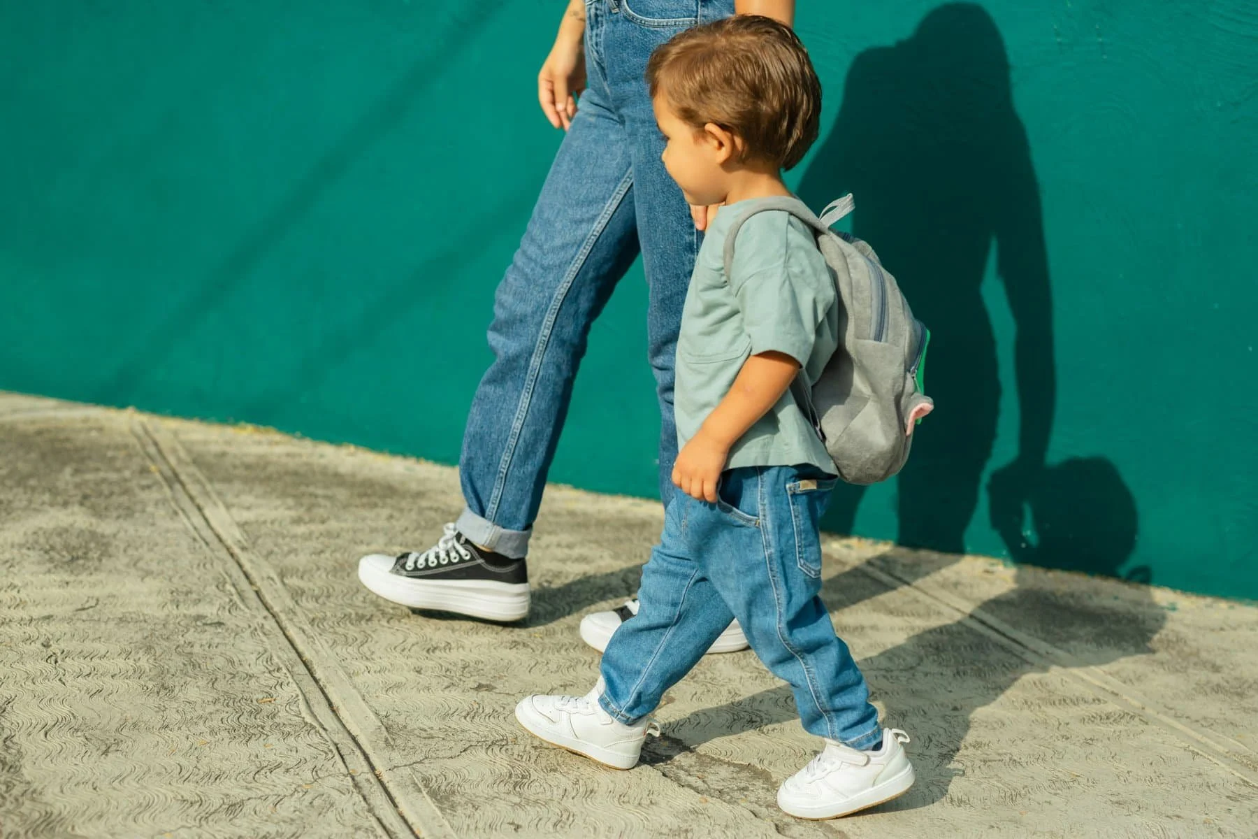 A young boy with a backpack walking on a sidewalk next to an adult. The boy is wearing white sneakers, blue jeans, a light green shirt, and has light brown hair. The adult is wearing black and white sneakers, blue jeans, and a green top. The background features a teal wall and sunlight casting shadows.