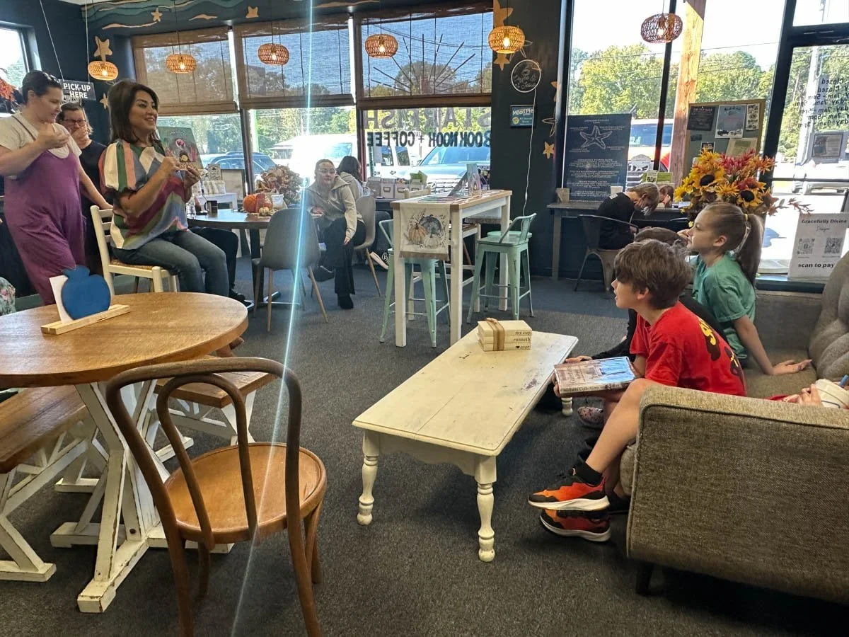 People sitting and standing inside a coffee shop, some children watching a woman speaking, decorated with hanging lamps, flowers, and signs about ordering and pickup.