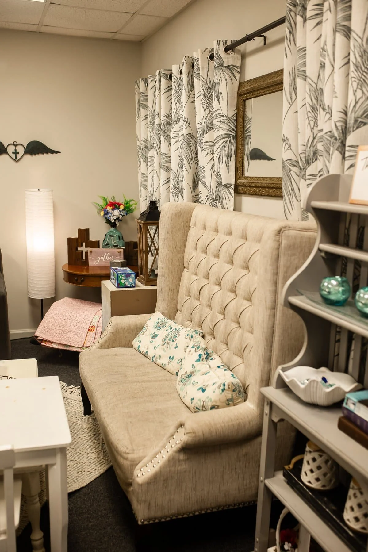 A cozy living room corner with a beige tufted high-back sofa, a decorative throw pillow, a small white side table, a patterned blanket on the floor, the wall behind has white curtains with black leaf pattern, a framed mirror, a small shelf with decorative glass balls, a floral arrangement, and wall decor with a heart, wings, and cross.
