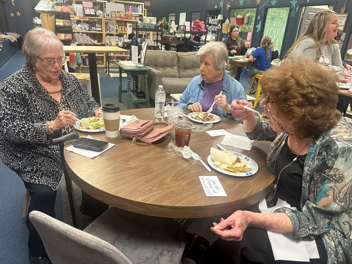 Three women eating lunch at a round table in a bookstore or library, with bookshelves and decor in the background.