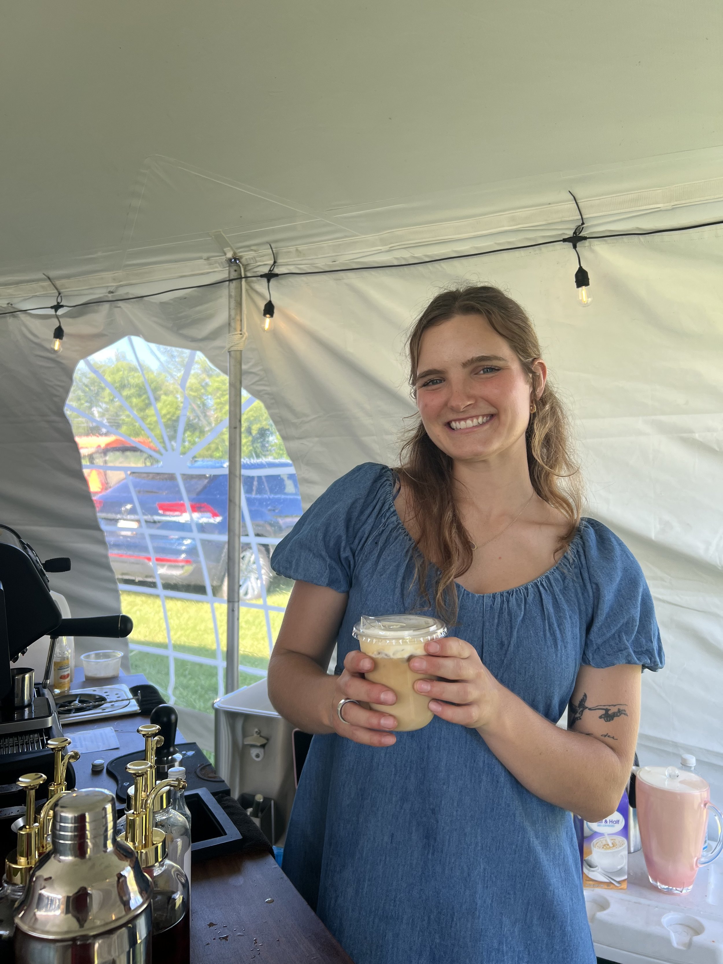 A young woman with long wavy hair and a tattoo on her arm, smiling and holding a glass of iced coffee inside a tent at an outdoor event. There are drink dispensers and a coffee machine on the counter in front of her.