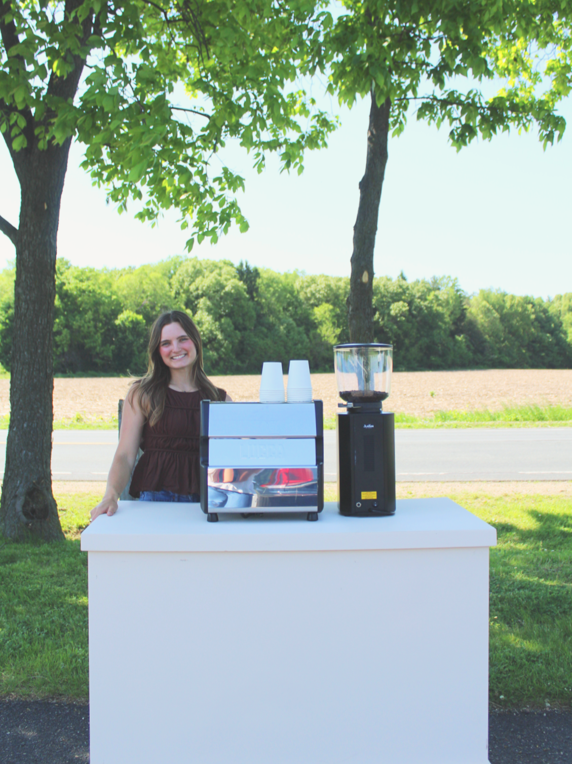 A woman smiling at a food stand with a coffee grinder and toaster outdoors on a white table, surrounded by trees and a field on a sunny day.