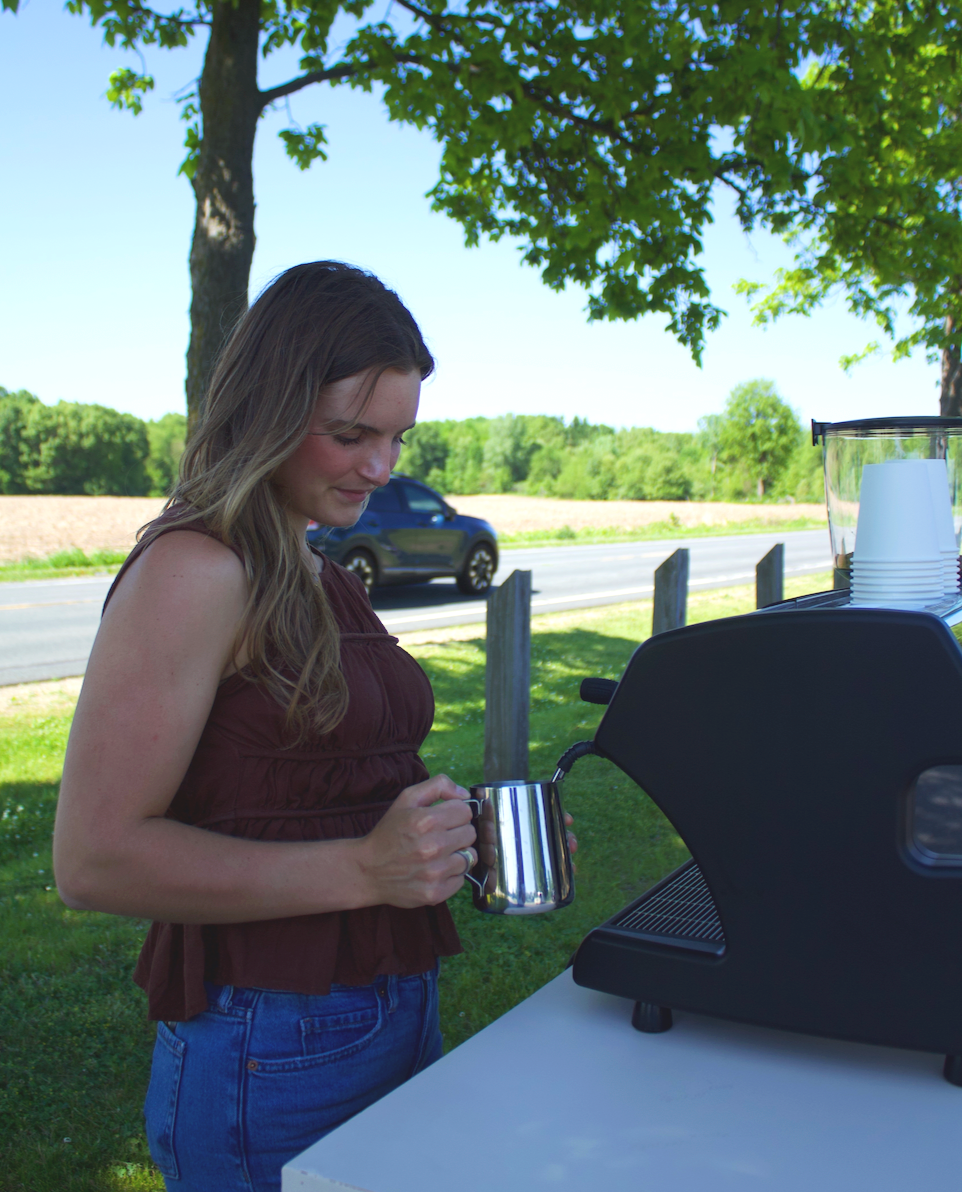 A woman preparing to make coffee using a commercial espresso machine outdoors under a tree on a sunny day.