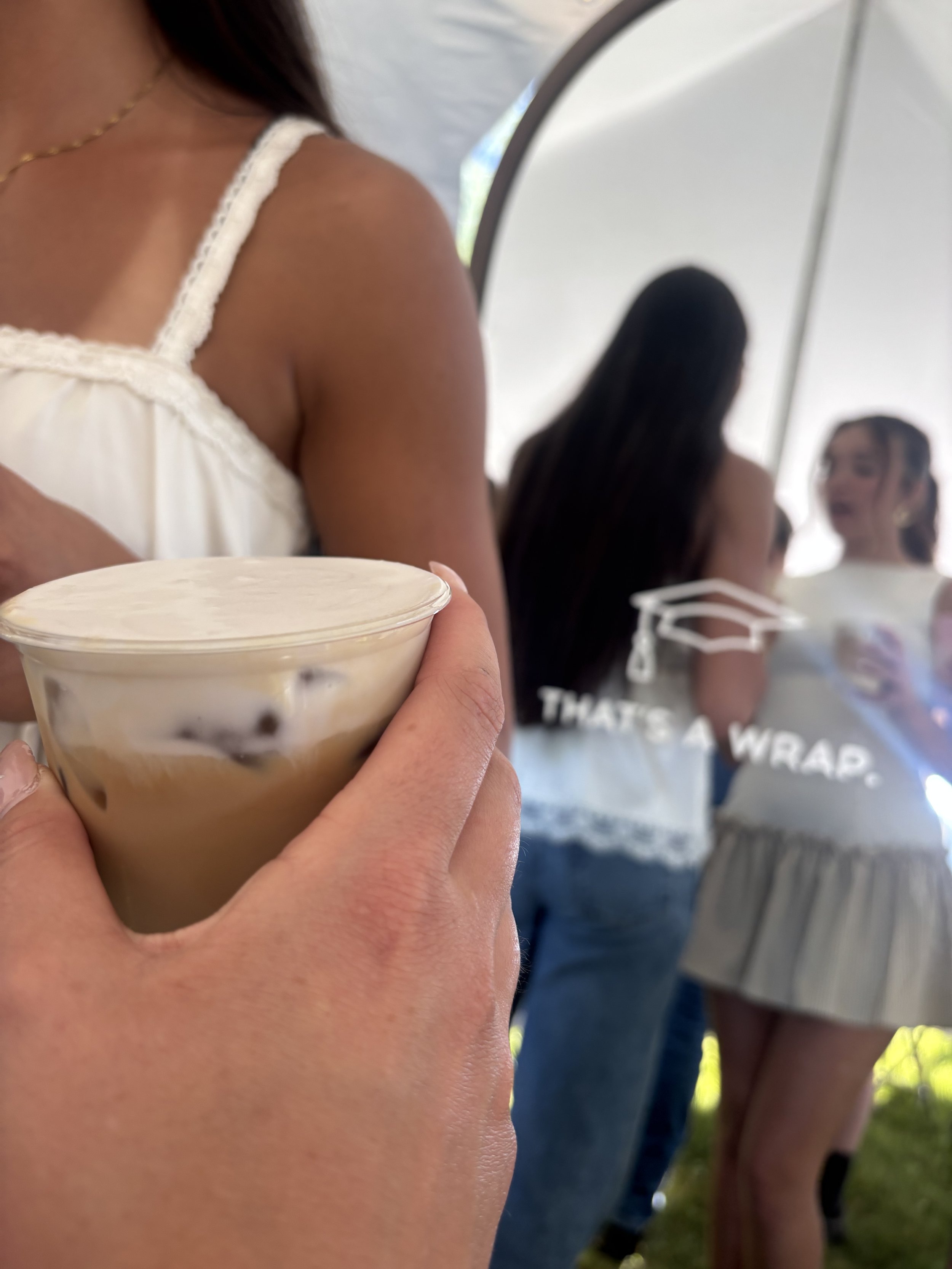 Person holding a cup of iced coffee or milk tea with tapioca pearls, with a woman in a white top and a group of people in the background.